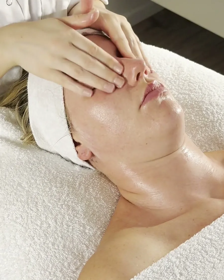 Two hands rest on the forehead of a client wearing a white headband, who is lying relaxed on the treatment bed