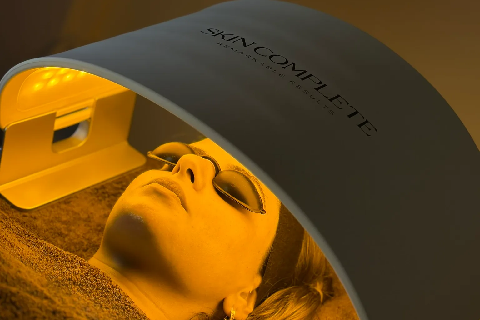 A client lies with covered eyes under the Skin Complete LED panel in the treatment room at Moons Beauty, in warm yellow light
