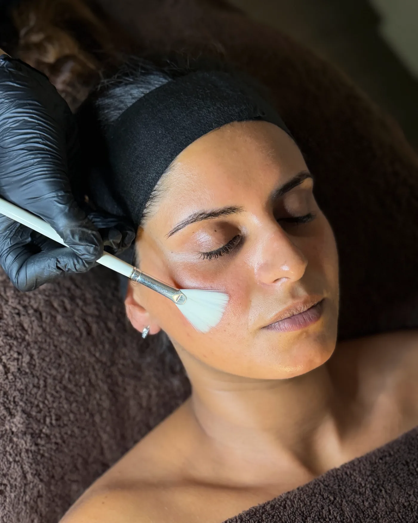A peel being applied with a fan brush to the cheek of a client wearing a black hairband
